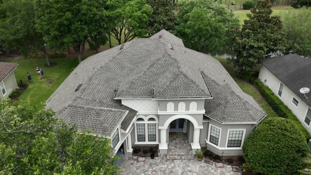 An aerial view of a large house with a gray roof, surrounded by greenery, with two people visible in the yard.