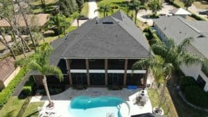 Aerial view of a two-story house with a shingled roof and a backyard swimming pool, surrounded by a fence, palm trees, and neatly landscaped lawns.