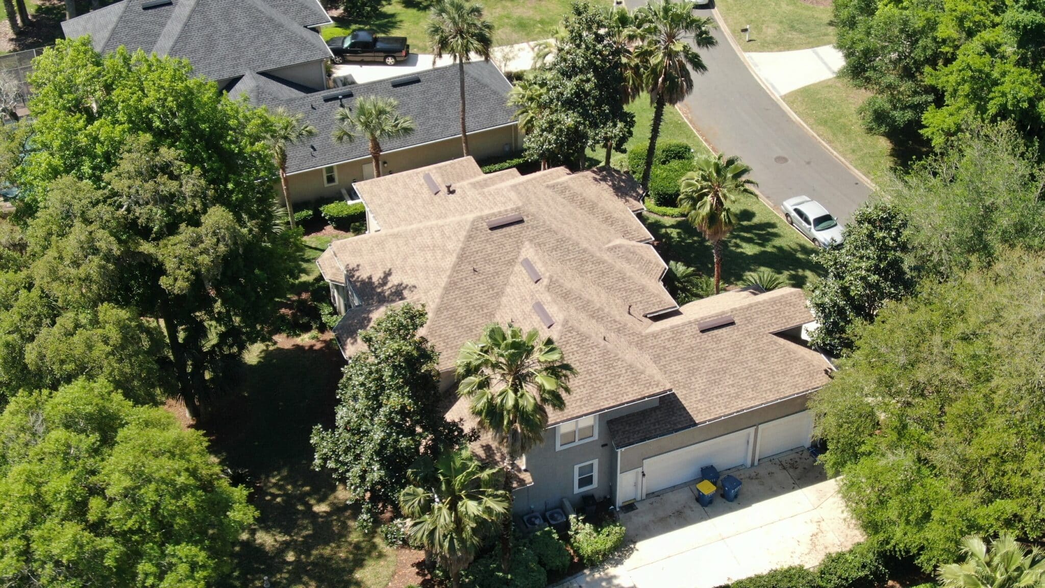 Aerial view of a suburban house with a shingled roof, surrounded by green trees, next to a road with a parked car.