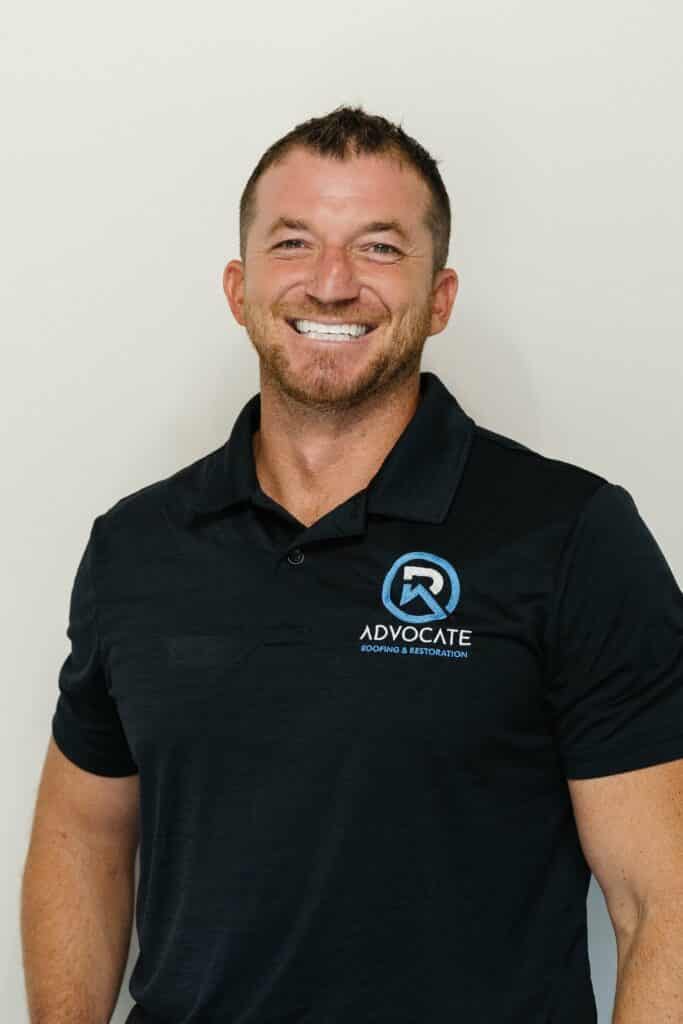 A smiling person with short brown hair, wearing a black polo shirt with a logo, stands against a light background. They have a friendly demeanor.