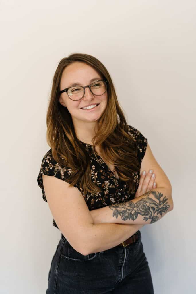 A person with glasses and a floral tattoo on their arm is smiling, wearing jeans and a flower-patterned shirt, standing against a white background.