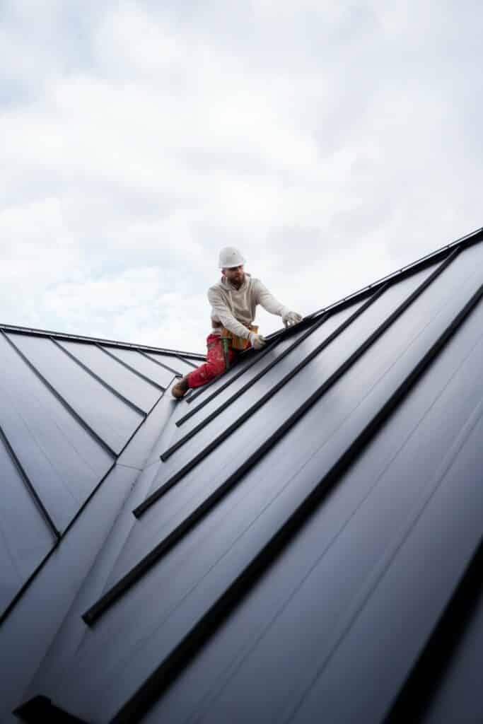 A person in a hardhat and gloves works atop a sloped, metallic roof structure against a cloudy sky backdrop, emphasizing construction and maintenance.