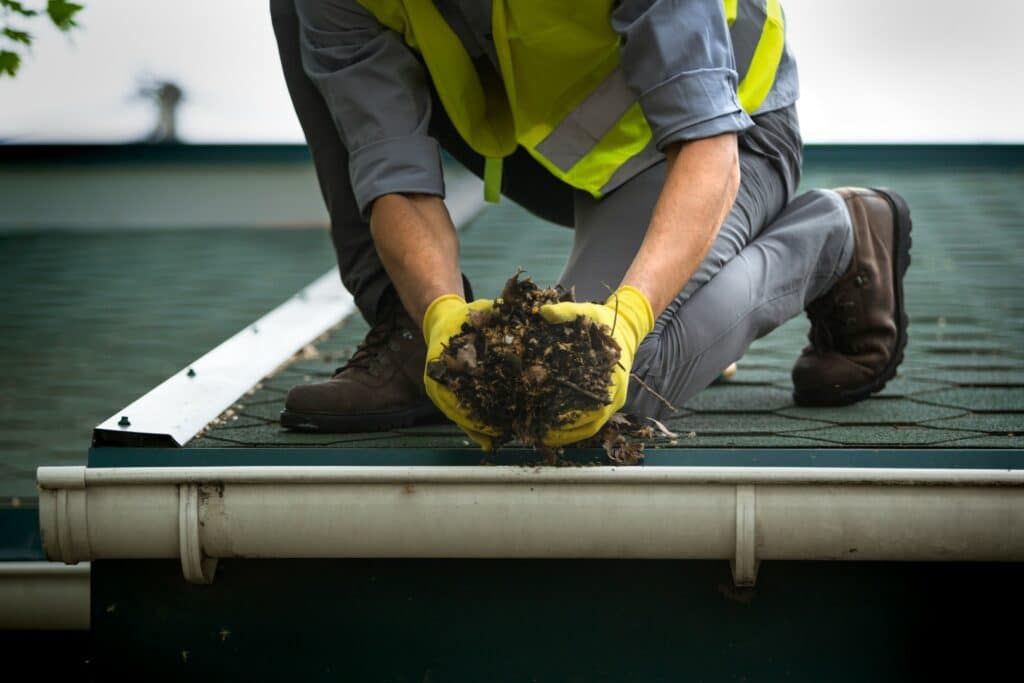 A person wearing a reflective vest and gloves is cleaning leaves from a gutter on a rooftop, with trees and overcast sky in the background.