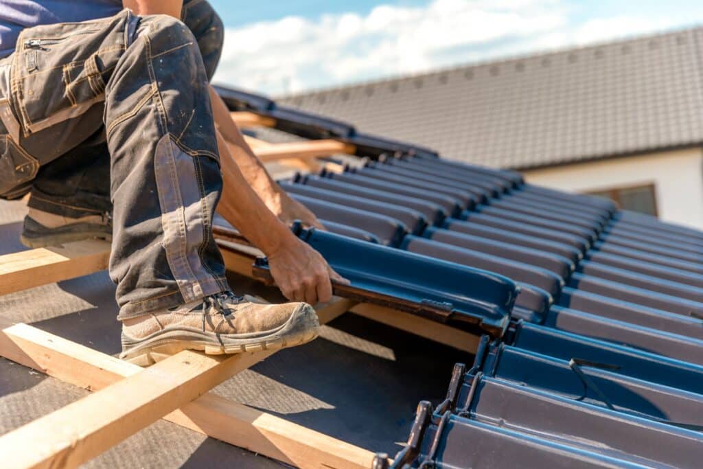 A person is seated on a rooftop installing dark tiles. The focus is on their hands and work boots, showcasing a sunny day with construction in progress.