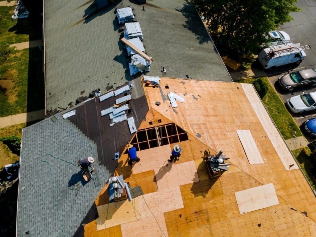 An aerial view of a residential building showing people working on roof construction, partially completed with shingles and wooden sheathing visible.