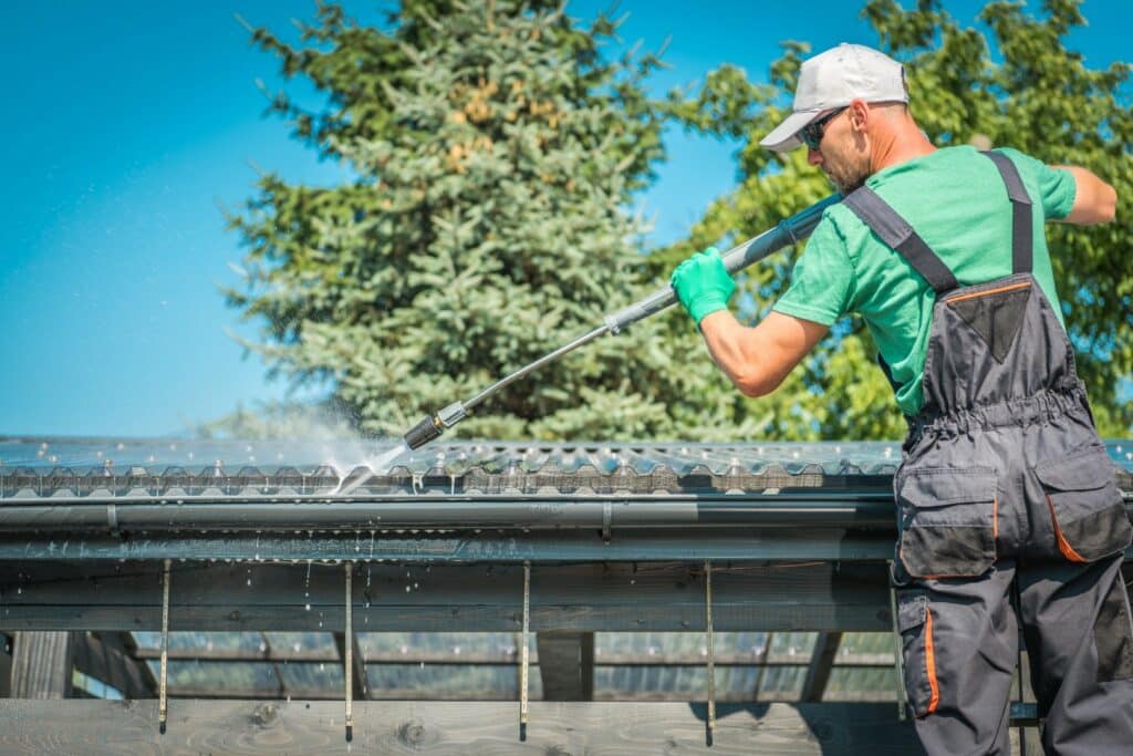 A person in a green shirt and protective gloves uses a high-pressure washer to clean a house's gutter under a clear blue sky.