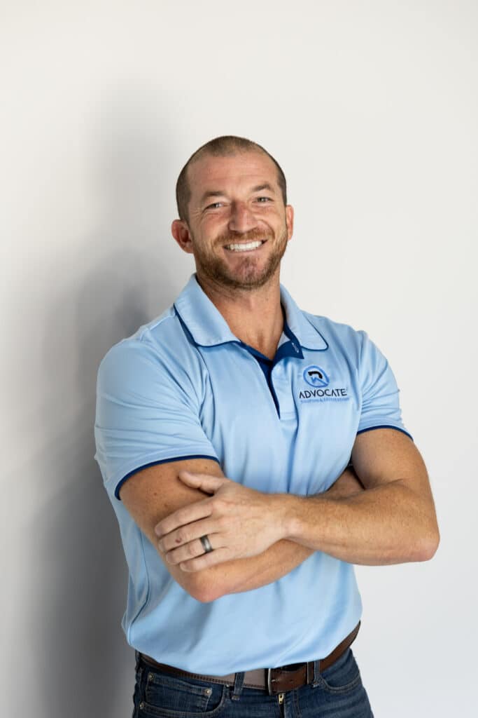 A smiling person with crossed arms wears a light blue polo shirt with a logo, standing confidently against a white background.