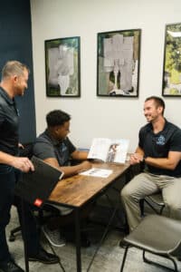 Three people in a modern office setting, smiling and engaging in a conversation with documents on a wooden table, art on the wall behind.